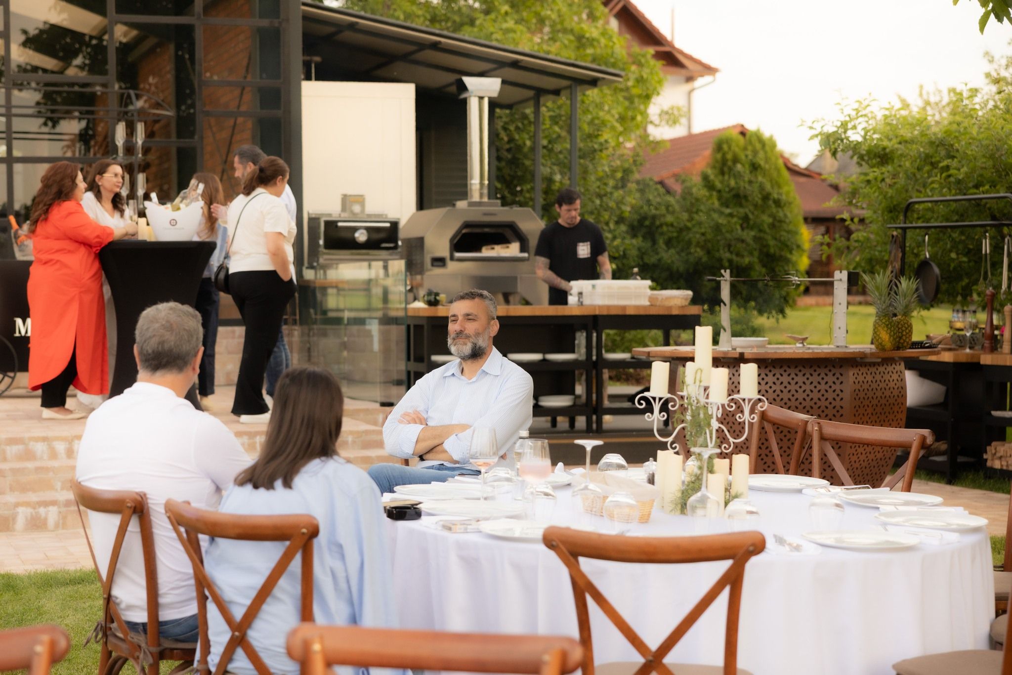 Masă lungă de eveniment, în aer liber, cu invitați așezați printre arbori